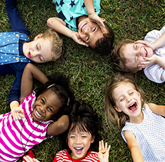 . Image - Six smiling children lying on grass with heads in the center. Click tile image to view